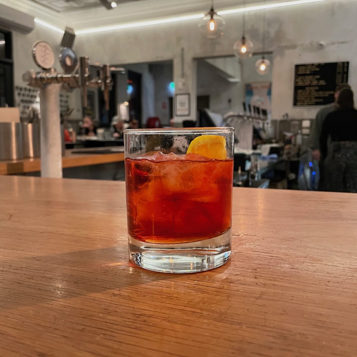 Glass with dark red liquid and a slice of orange on a light wooden bench